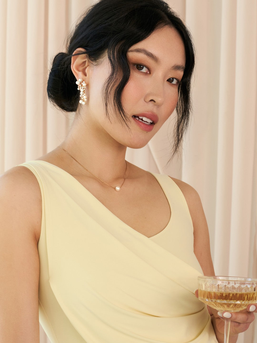 Woman in a light yellow dress with earrings holding a glass of champagne against a neutral background