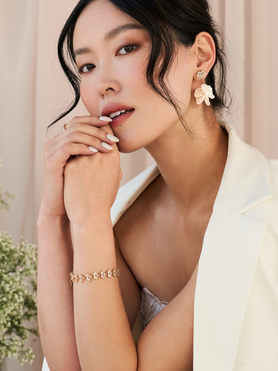 Woman wearing a white outfit with floral earrings and a gold bracelet against a light background