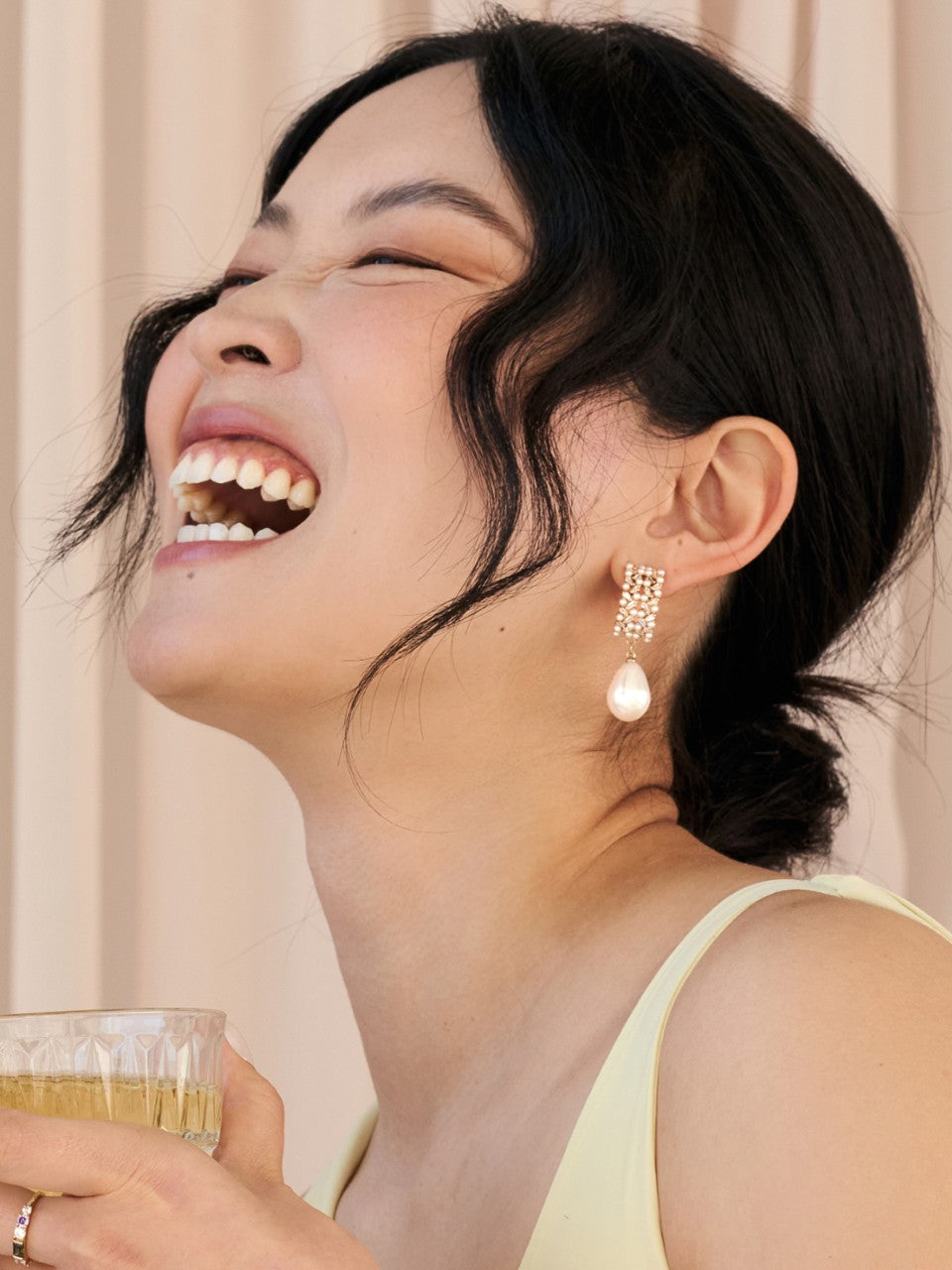 Woman laughing with a glass of wine, wearing earrings and a light top.